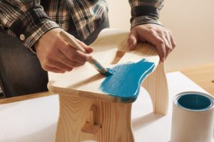 Craftsman varnishing a wooden handmade stool at home with a blue coating on a work table, hands close up